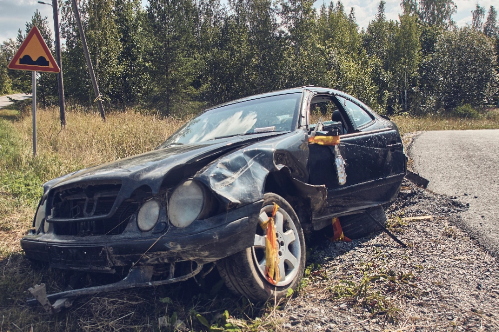 bmw-crashed-on-road-side A damaged black car crashed on the roadside next to an uneven road sign in a rural setting with trees and a clear sky, depicting an incident potentially related to running red lights.